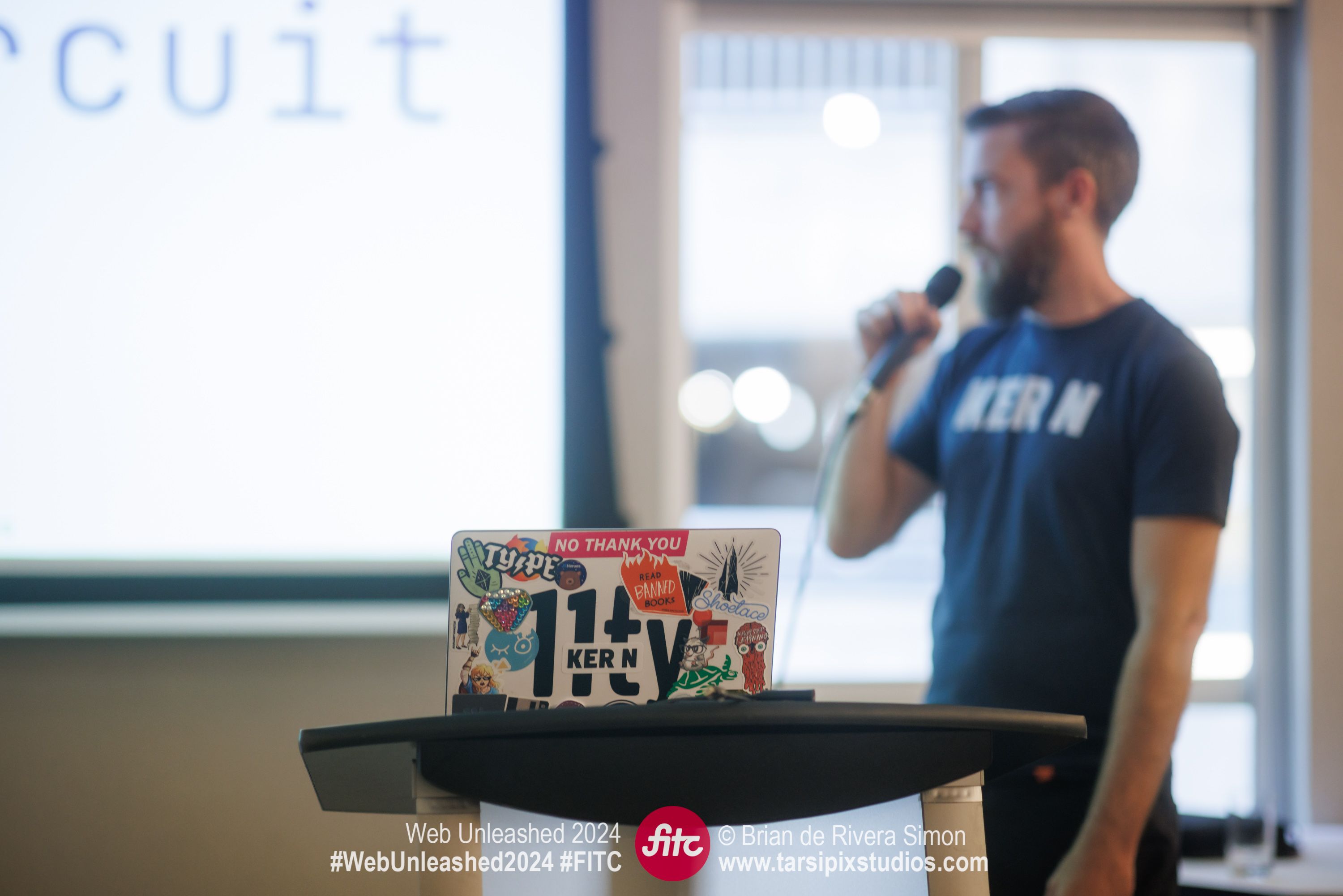 A laptop sits in focus on a podium. A blurred Zach stands in the background holding a microphone.